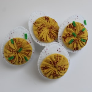 Pode incluir: Quatro ovos de feltro recheados em pratos de feltro branco. Os ovos são amarelos com detalhes em feltro vermelho e verde.
