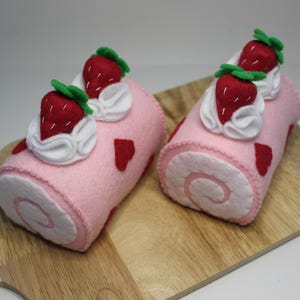 May include: Two pink felt strawberry roll cakes with red heart accents and white cream topping with red strawberries and green leaves, displayed on a wooden cutting board. The cakes have a spiral design.