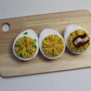 Puede incluir: Tres huevos rellenos de fieltro sobre una tabla de cortar de madera. Los huevos están decorados con fieltro amarillo, rojo y verde. Un huevo tiene una tira de fieltro marrón en la parte superior.