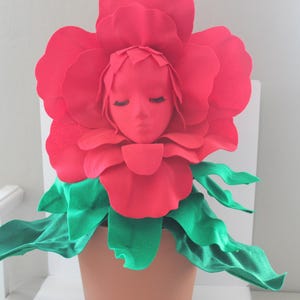 May include: A red felt flower with a human face in the center. The flower is in a brown pot with green felt leaves.
