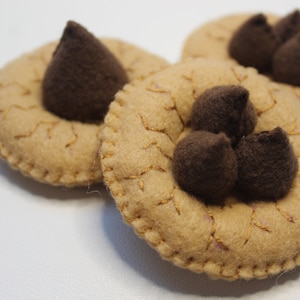 May include: Close-up of three felt cookies with brown chocolate chip accents. The cookies are tan with gold stitching and a stitched border. The chocolate chips are dark brown and cone-shaped. The cookies are arranged on a white surface.