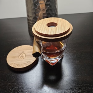Puede incluir: Un posavasos de madera con la palabra "Smoker" grabada, un vaso con un líquido marrón y una tapa de madera con un agujero en el centro. El vaso está sobre el posavasos y la tapa está sobre el vaso.
