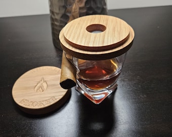 May include: A wooden coaster with the word "Smoker" engraved on it, a glass with a brown liquid, and a wooden lid with a hole in the center. The glass is sitting on top of the coaster, and the lid is on top of the glass.