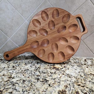 May include: A wooden egg tray with a handle. The tray has 18 individual indentations for holding eggs. The tray is made of light brown wood and has a natural finish.