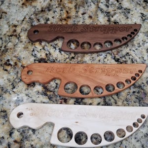 May include: Three wooden herb strippers, one in light wood, one in cherry wood, and one in walnut wood. Each stripper has a handle and a row of holes for stripping herbs.