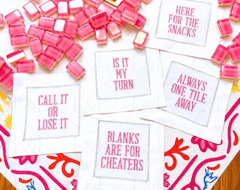 May include: Four white square fabric napkins with pink embroidered text reading HERE FOR THE SNACKS, IS IT MY TURN, ALWAYS ONE TILE AWAY, CALL IT OR LOSE IT, and BLANKS ARE FOR CHEATERS, surrounded by scattered pink and white game tiles.