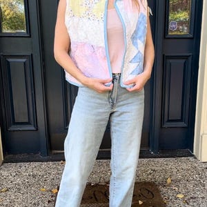 May include: A sleeveless quilted vest with a patchwork design in shades of pink, blue, and yellow. The vest has a light blue trim and is worn over a pink tank top.