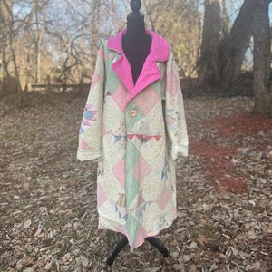 May include: A long, quilted coat with a patchwork design in pastel colors of pink, green, and cream. The coat features a bright pink collar and lining, with a single wooden button closure. The coat is displayed on a black mannequin.