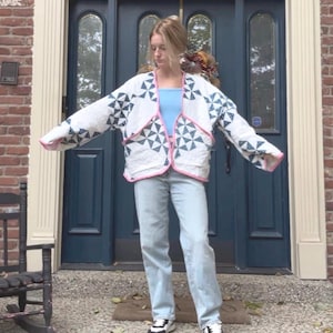 May include: A white quilted jacket with a geometric blue and white pattern, trimmed with pink. The jacket is worn over a light blue top and paired with light wash jeans and sneakers. The jacket's design features a repeating pattern of blue triangles.
