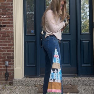 May include: Blue denim jeans with a patchwork design on the side. The patchwork includes various fabrics with floral, polka dot, and plaid patterns. The jeans are worn with a light pink sweater.