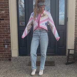 May include: A cropped, quilted jacket with a pink and white design, paired with light wash jeans and white boots. The jacket features a patchwork pattern with floral and geometric elements. The outfit is completed with a pink top and a gray headband.