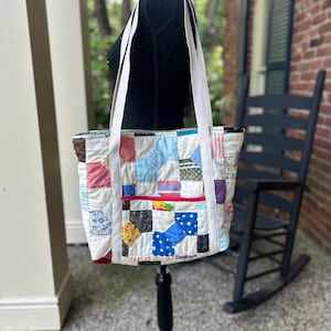 May include: A patchwork tote bag with white straps. The bag is made of various fabric squares in colors like blue, red, yellow, and green. A red zipper pocket is visible on the front. The bag is displayed on a black mannequin.