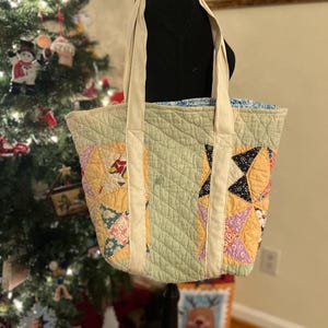 May include: A quilted tote bag with cream-colored handles. The bag features a patchwork design with various fabric patterns in shades of yellow, pink, blue, and black. The main body of the bag is a light green color.