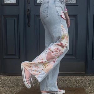 May include: Light wash denim jeans with floral and heart patchwork detail on the side. The jeans are paired with pink sneakers. The patchwork features a variety of floral patterns and a pink heart.