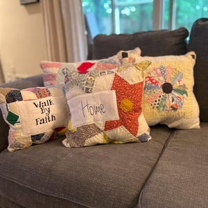 May include: A collection of handmade, patchwork pillows in various colors and patterns. One pillow reads "Walk by Faith," and another says "Home." The pillows are arranged on a gray sofa, creating a cozy and inviting atmosphere.