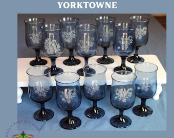 May include: A set of Pfaltzgraff Yorktowne water goblets. The blue-tinted glasses have a floral design and a dark blue stem. The words "WATER GOBLETS" are at the bottom of the image.