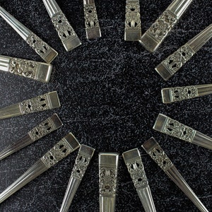 May include: Silverware set arranged in a circular pattern. The utensils have ornate, detailed handles with a floral design. The polished metal reflects light, creating a contrast against the dark background. A classic set for dining.