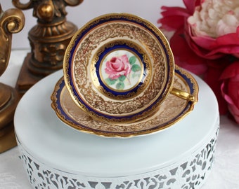 May include: A vintage teacup and saucer with a gold and blue floral design. The cup features a pink rose with green leaves on a blue background. The saucer has a gold rim and a blue and gold floral pattern.