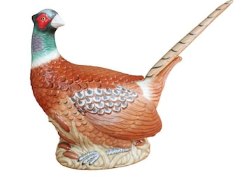 May include: A ceramic pheasant figurine with detailed feather textures in shades of brown, orange, and white. The bird has a red face, green neck, and a long, striped tail. It is perched on a base of straw-like material.