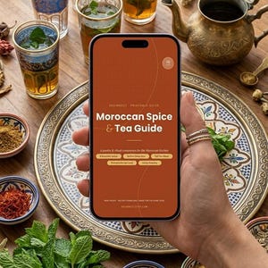 May include: A smartphone displaying a digital Moroccan Spice & Tea Guide with orange background and white text. The screen shows the title, subheadings, and small icons. Surrounding the phone are small bowls of spices, mint, and tea glasses.