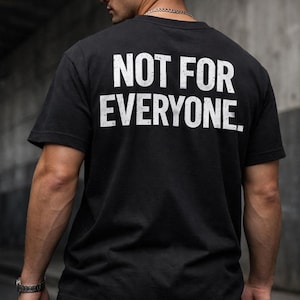 Op de afbeelding: Zwart T-shirt met de witte tekst "NOT FOR EVERYONE." Het shirt met korte mouwen lijkt van een zacht materiaal te zijn. De persoon op de foto draagt een zwarte baseball cap en een zilveren ketting.