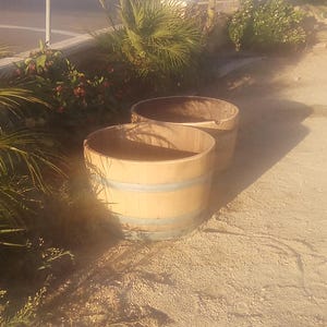 May include: Two light-colored wooden barrels with smooth staves and two horizontal metal bands each. The barrels are open at the top and have a cylindrical shape, positioned side-by-side on a light-colored, textured ground surface.