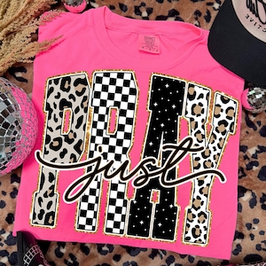 Peut inclure: Un t-shirt rose vif avec l'inscription "PRAY Just" dans une police audacieuse et stylisée. Les lettres présentent des motifs léopard, à carreaux et étoilés. Une casquette de baseball noire et blanche et des lunettes de soleil sont également visibles.