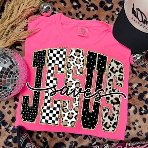 May include: A vibrant pink t-shirt with the text "JESUS SAVES" in a decorative font. The letters are a mix of black and white checkered, leopard print, and gold. A black and white baseball cap is also visible in the image.