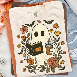 May include: A cream-coloured t-shirt featuring a graphic of a ghost reading a book with a pumpkin on the cover while holding an iced coffee. The ghost is surrounded by autumn-themed illustrations including orange flowers, brown leaves, and black bats.