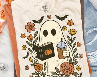 May include: A cream-colored t-shirt featuring a graphic of a ghost reading a book with a pumpkin on the cover while holding an iced coffee. The ghost is surrounded by autumn-themed illustrations including orange flowers, brown leaves, and black bats.