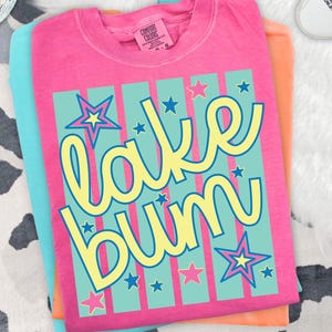 Op de afbeelding: Een stapel van drie t-shirts in roze, turquoise en oranje. Het bovenste shirt is roze met de tekst "lake bum" in gele letters op een turquoise gestreepte achtergrond met sterren. De shirts zijn netjes opgevouwen.