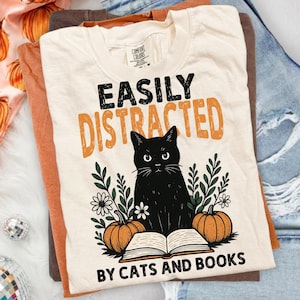 May include: A cream-colored t-shirt featuring a black cat sitting behind an open book, flanked by two orange pumpkins and green foliage. The text reads EASILY DISTRACTED BY CATS AND BOOKS in black and orange lettering on the front of the garment.