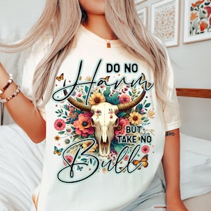 Może przedstawiać: Kremowa koszulka z motywem czaszki krowy z kwiatowymi akcentami i napisem "DO NO HARM BUT TAKE NO BULL". Projekt zawiera motyle i kolorowe kwiaty. Swobodny t-shirt z grafiką.