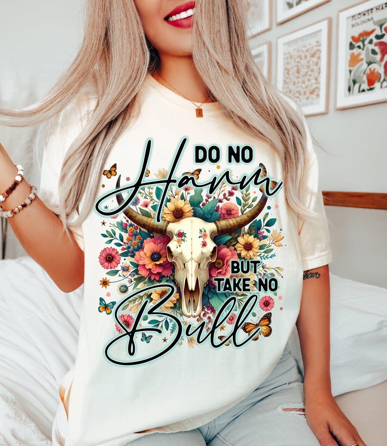 Może przedstawiać: Kremowa koszulka z motywem czaszki krowy z kwiatowymi akcentami i napisem "DO NO HARM BUT TAKE NO BULL". Projekt zawiera motyle i kolorowe kwiaty. Swobodny t-shirt z grafiką.