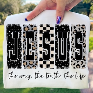 Könnte beinhalten: Weißes T-Shirt mit dem Wort "JESUS" in einem kräftigen Blockbuchstaben-Design. Jeder Buchstabe hat ein einzigartiges Muster, darunter Polka Dots, Leopardenmuster und ein Schachbrettmuster. Der Satz "the way, the truth, the life" steht darunter.