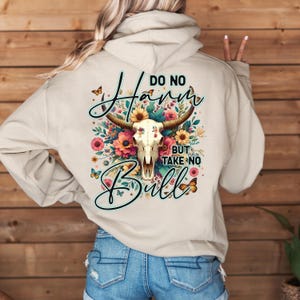 Może przedstawiać: Beżowa bluza z kapturem z grafiką. Projekt przedstawia czaszkę byka, kwiaty i napis "Do No Harm But Take No Bull". Projekt zawiera motyle i skrypty. Bluza wykonana jest z miękkiego materiału.