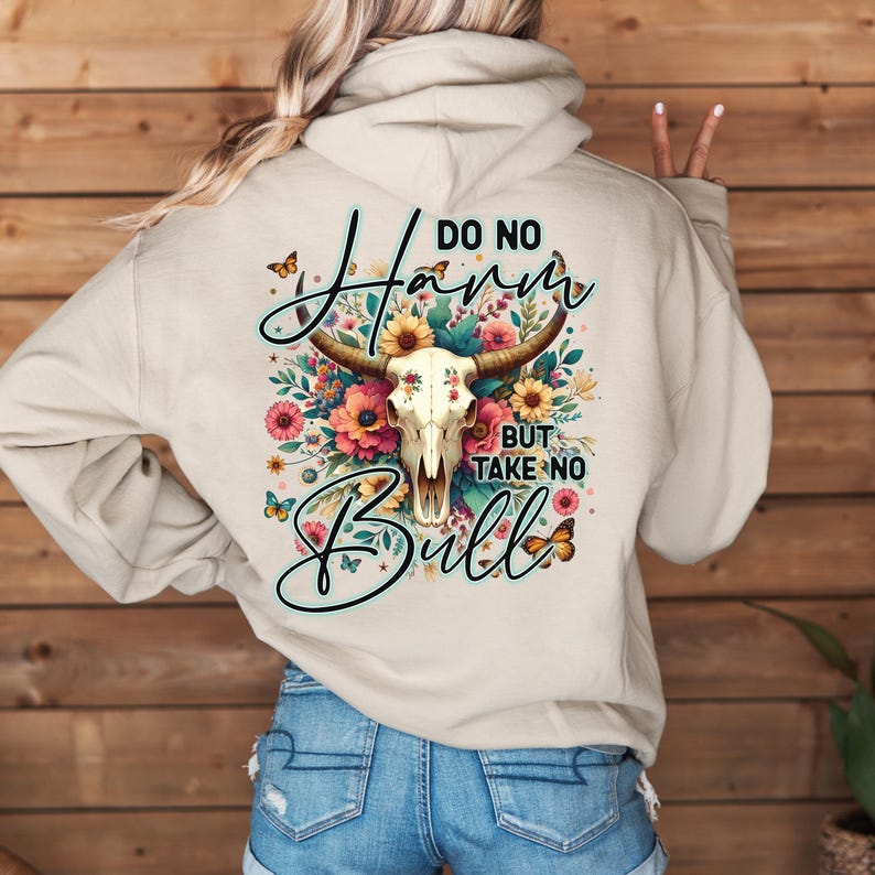 Może przedstawiać: Beżowa bluza z kapturem z grafiką. Projekt przedstawia czaszkę byka, kwiaty i napis "Do No Harm But Take No Bull". Projekt zawiera motyle i skrypty. Bluza wykonana jest z miękkiego materiału.