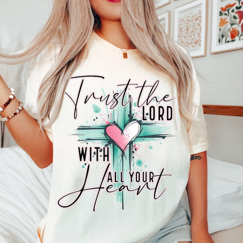 May include: A white t-shirt with a teal and pink graphic of a cross with a heart in the center. The text "Trust the Lord with all your heart" is written on the shirt.