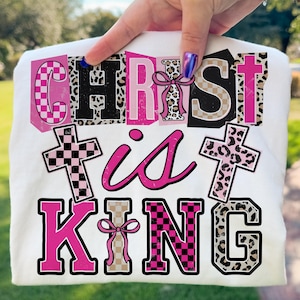 May include: White t-shirt with the words "CHRIST is KING" in a mix of pink, black, and leopard print letters. The design includes cross graphics and a bow.