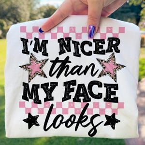 Könnte beinhalten: Weißes T-Shirt mit dem Text "I'M NICER than MY FACE looks" in Schwarz, mit rosa und Leopardenmuster Sternakzenten und einem rosa Karomuster. Das T-Shirt wird von einer Hand gehalten.