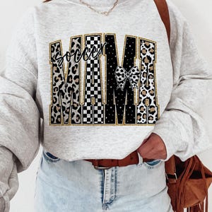May include: A light gray sweatshirt with the words "Soccer Mama" in a decorative design. The word "Soccer" is in a script font, and "Mama" is in a bold, block letter style. The letters have a mix of leopard print, black and white checkered patterns, and a soccer ball bow.