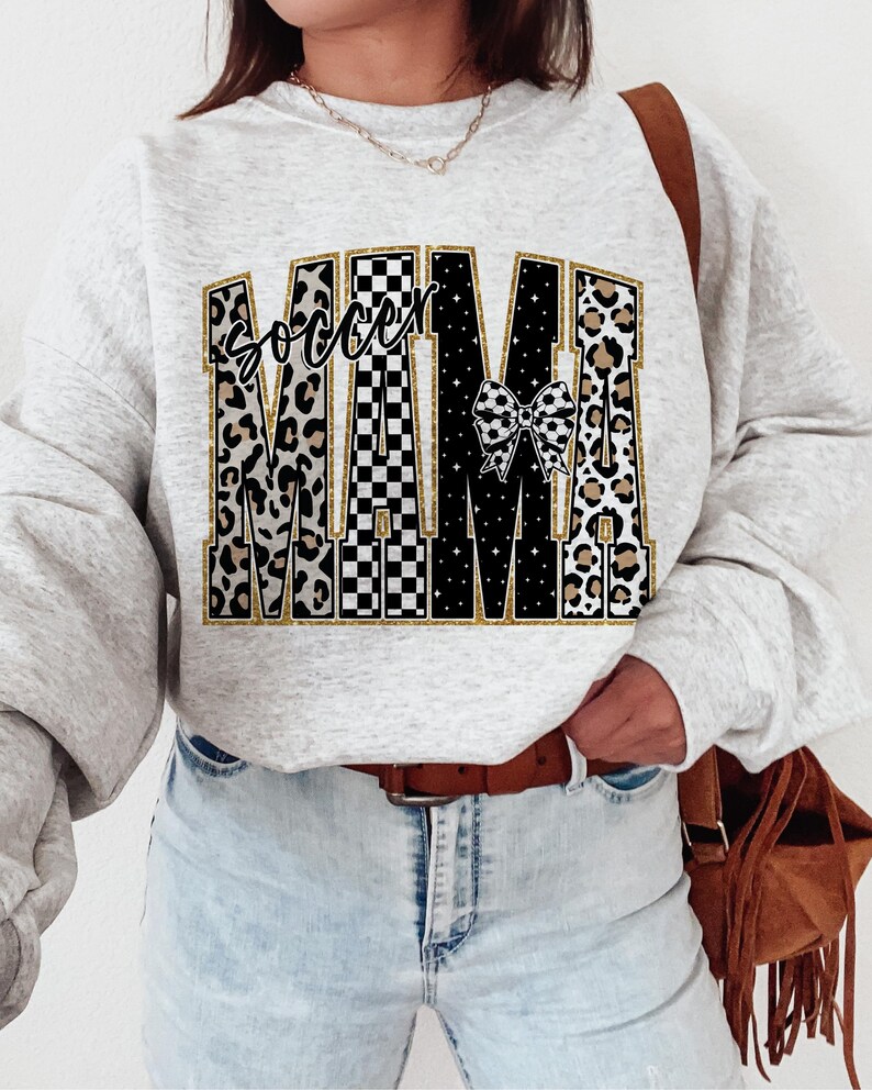 May include: A light gray sweatshirt with the words "Soccer Mama" in a decorative design. The word "Soccer" is in a script font, and "Mama" is in a bold, block letter style. The letters have a mix of leopard print, black and white checkered patterns, and a soccer ball bow.