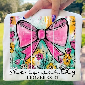 May include: White t-shirt featuring a vibrant, artistic design. A large pink bow is the focal point, set against a backdrop of colorful brushstrokes and floral accents. The text "She is worthy PROVERBS 31" is written in a stylish script.