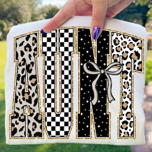 May include: White t-shirt with the word "AUNT" in large, bold letters. The letters have a gold glitter outline and are filled with leopard print, a black and white checkerboard pattern, and a black pattern with white stars. A bow is in the center.