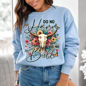 Może przedstawiać: Jasnoniebieska bluza z napisem "Do No Harm But Take No Bull". Projekt przedstawia czaszkę byka z kwiatowymi akcentami w odcieniach r&oacute;żu, ż&oacute;łci i pomarańczy. Motyle są rozrzucone po całym projekcie.
