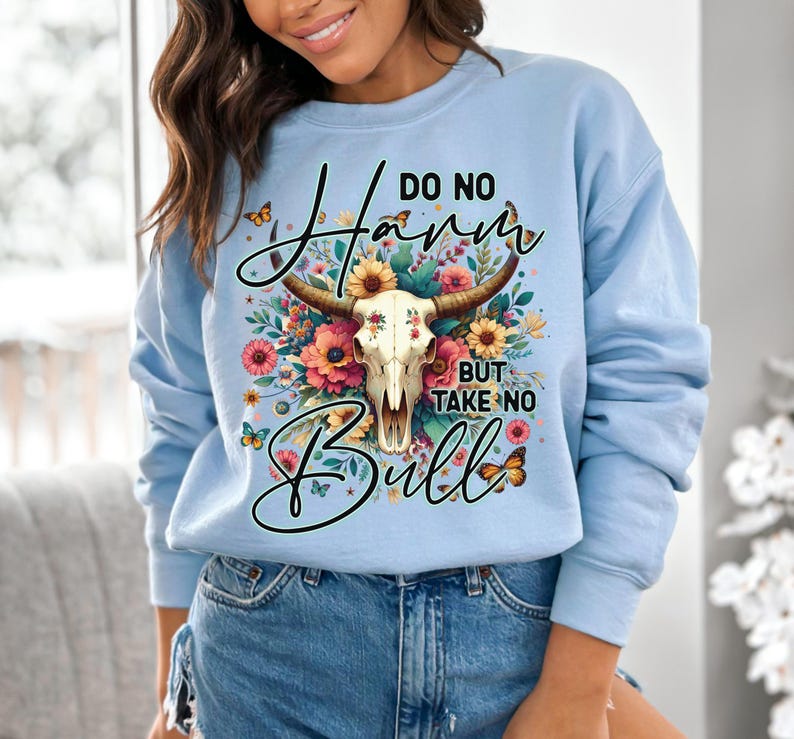 Może przedstawiać: Jasnoniebieska bluza z napisem "Do No Harm But Take No Bull". Projekt przedstawia czaszkę byka z kwiatowymi akcentami w odcieniach r&oacute;żu, ż&oacute;łci i pomarańczy. Motyle są rozrzucone po całym projekcie.