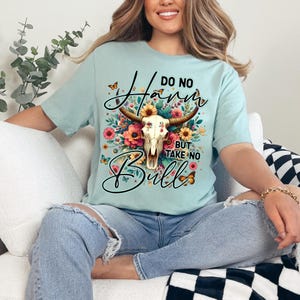 Może przedstawiać: Jasnoniebieska koszulka z motywem czaszki byka z kwiatowymi akcentami i napisem "Do No Harm But Take No Bull". Projekt zawiera motyle i kolorowe kwiaty.