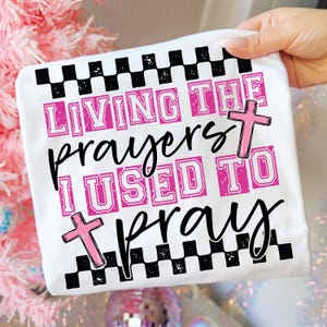May include: White t-shirt with the text "LIVING THE prayers I USED TO pray" in pink and black. The design includes two pink crosses and a black and white checkered pattern.