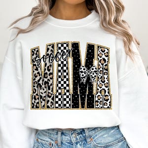 May include: White sweatshirt with the text "Soccer Mama" in a bold, decorative font. The letters have a mix of patterns, including leopard print, black and white checkerboard, and a soccer ball bow.