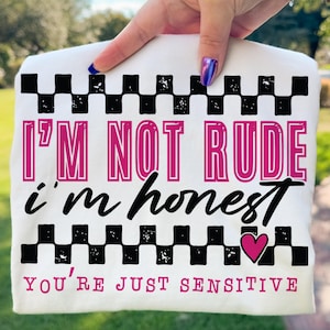 May include: White t-shirt with the text "I'M NOT RUDE i'm honest YOU'RE JUST SENSITIVE". The design includes a black and white checkered pattern and a pink heart.